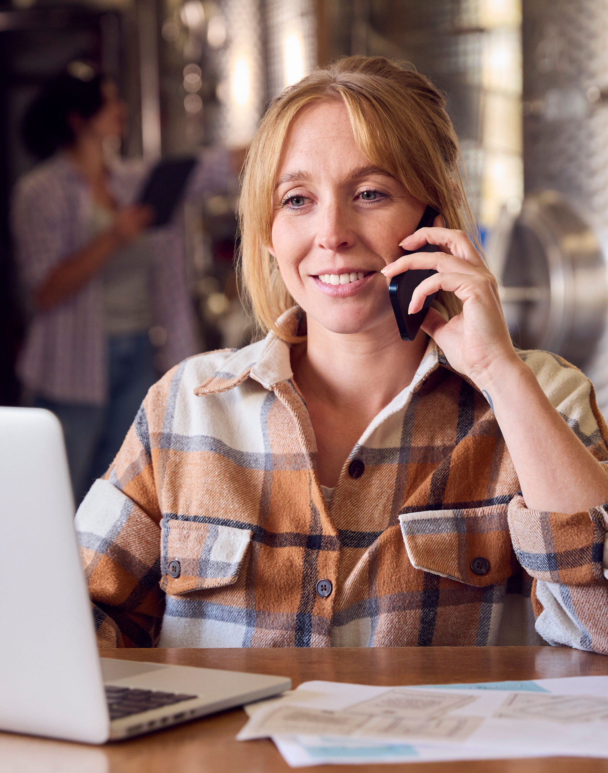 Woman On Mobile Phone With Laptop And Wine Bottles Inside Winery